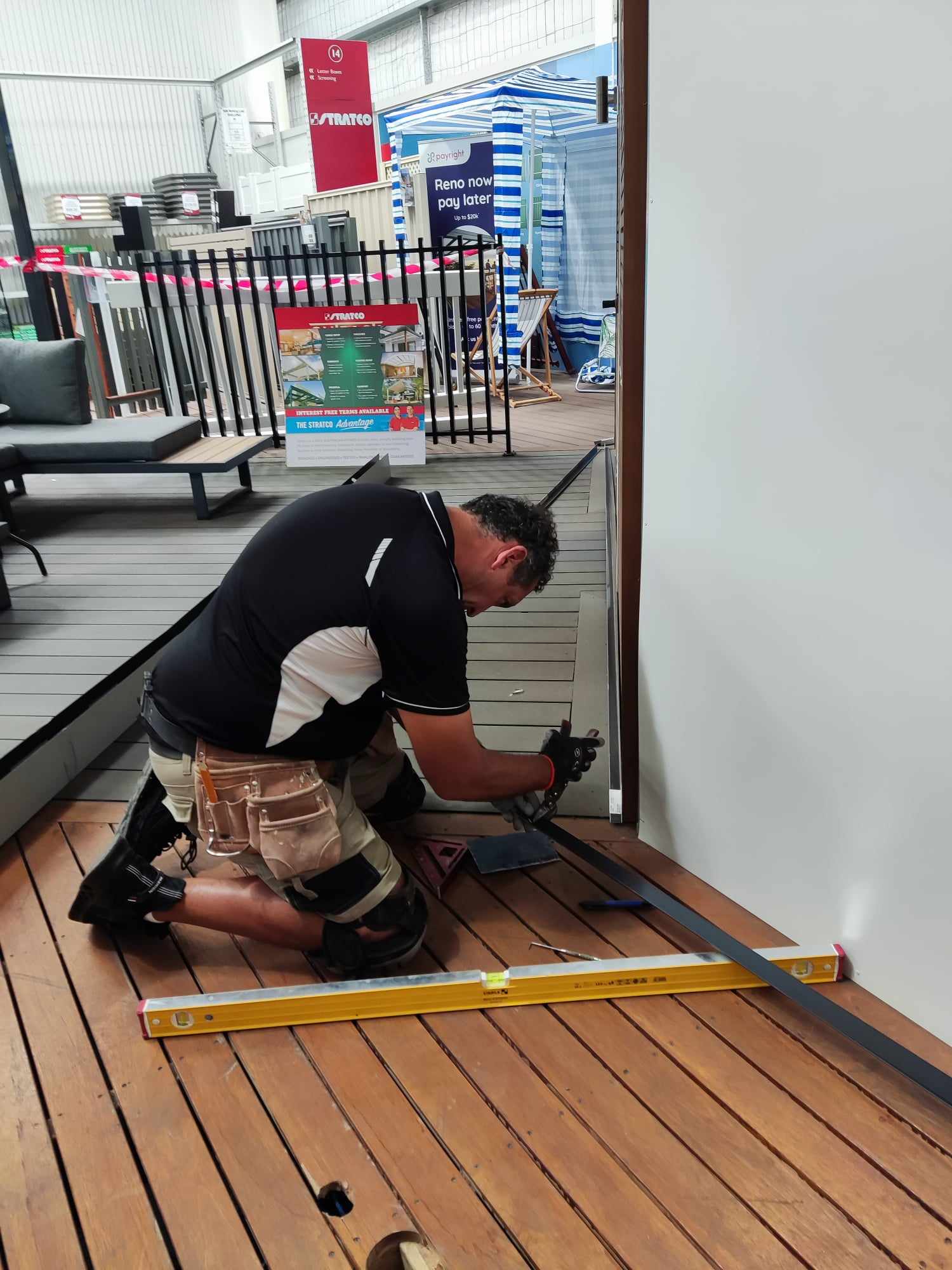 A man kneels on a wooden deck, using a tool to work on the floor near a white wall. Construction and Maintenance Solutions tools like a yellow level and measuring tape are on the deck, with display stands and banners in the background.