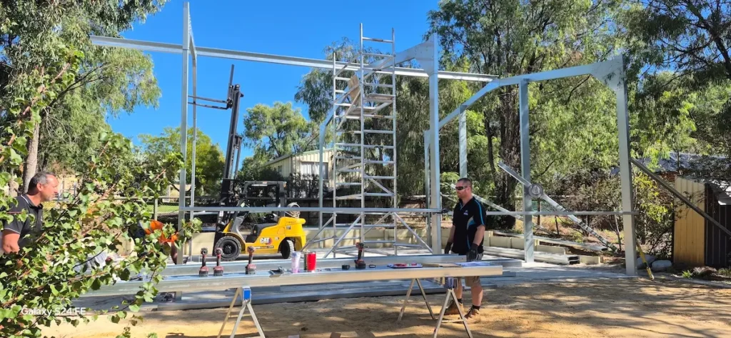Two men work at a construction site with a metal frame structure being assembled. Tools and materials are laid out on a table, and a yellow forklift is visible in the background among trees and buildings.