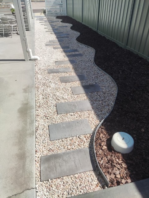 A walkway made of rectangular stepping stones set in gravel runs beside a wavy border separating dark mulch from light stones—expertly crafted with Construction and Maintenance Solutions—next to a metal fence and concrete patio.
