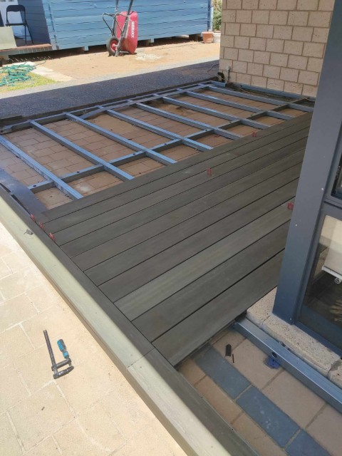 A partially constructed outdoor deck with metal framing and several wooden planks installed showcases quality Construction and Maintenance Solutions, next to a brick wall and glass door. Tools and construction materials are visible nearby.